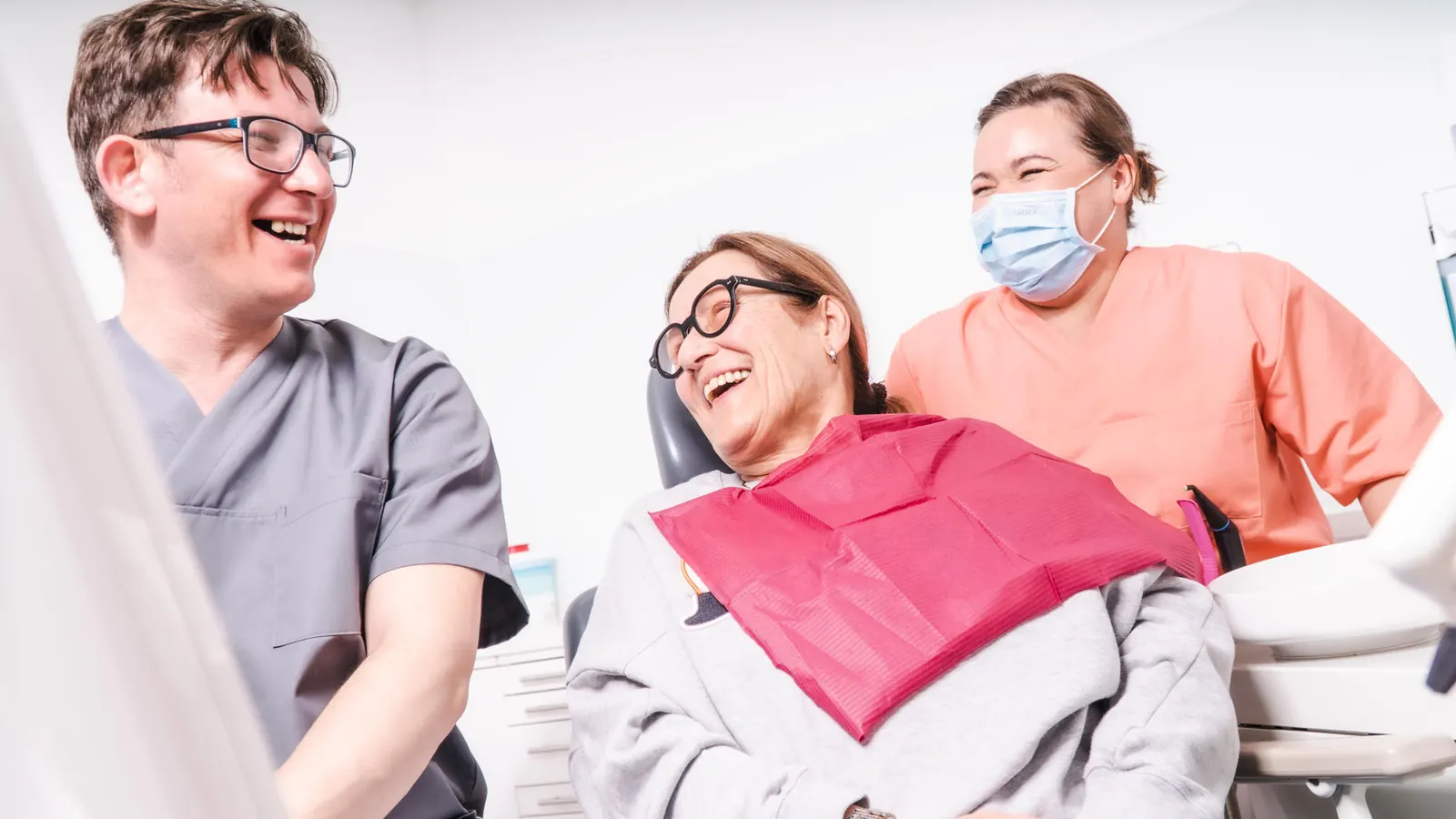Lachende Patientin im Behandlungsstuhl mit zwei Mitarbeitern der Praxis